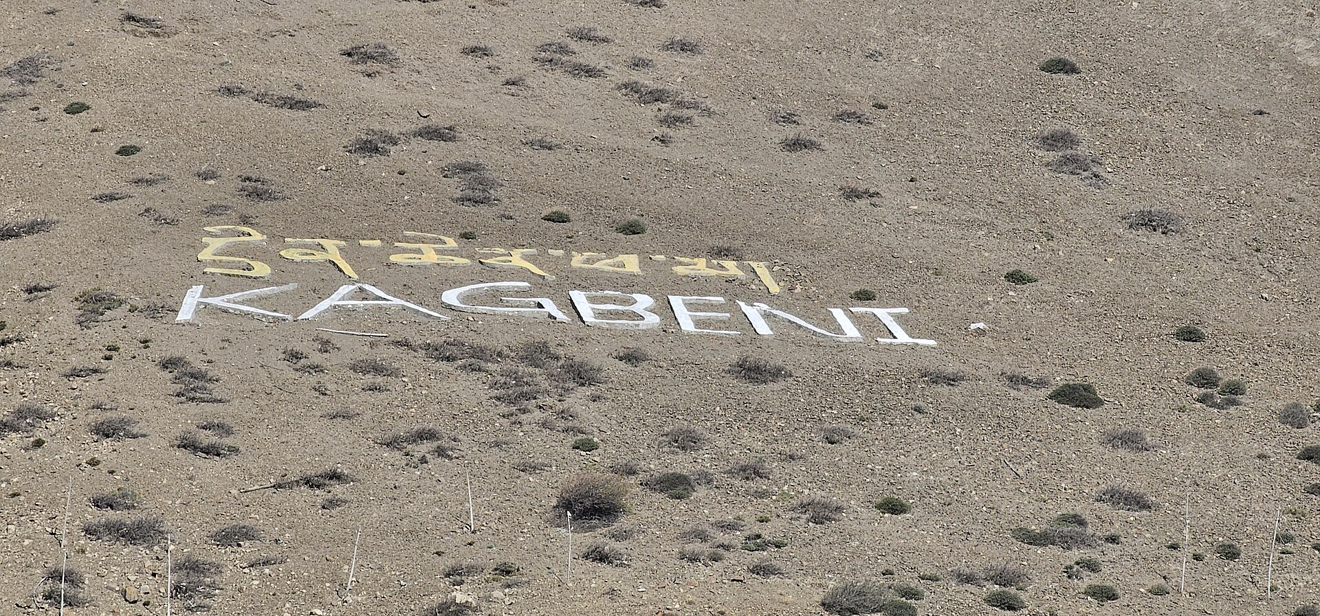 The word KAGBENI written in giant white letters on a stony hillside, with Tibetan script above