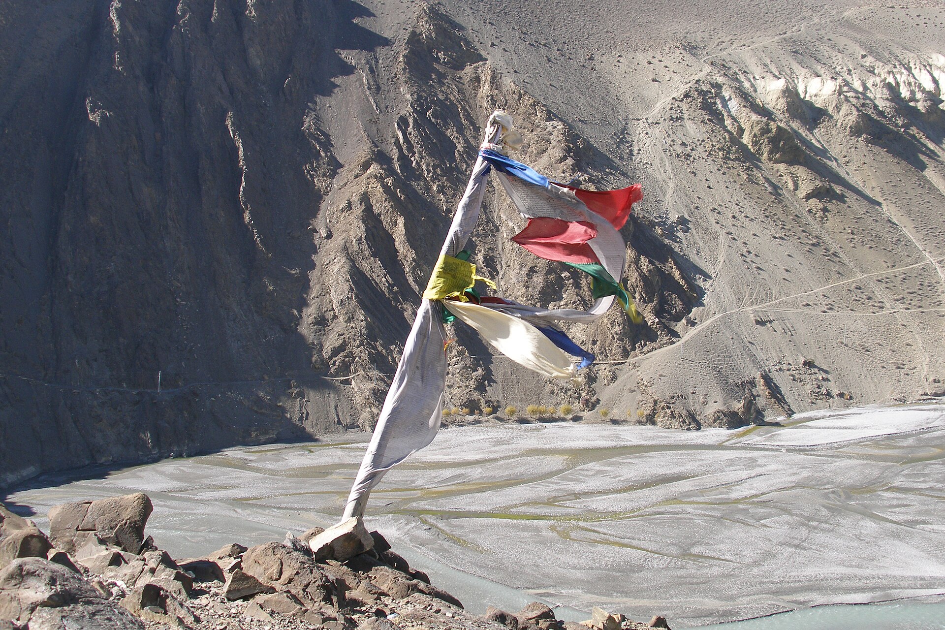 A tattered prayer flag whipping in strong wind above the wide gravel riverbed